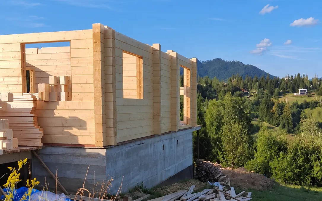 A log house under construction on a concrete base, with mountains behind